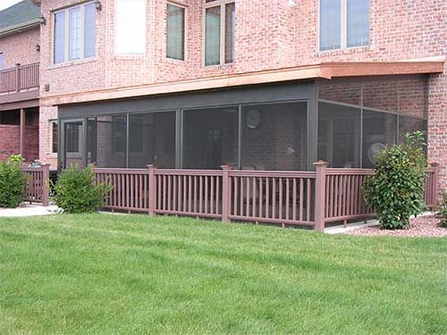 A screened in porch with a wooden railing and a brick house in the background.