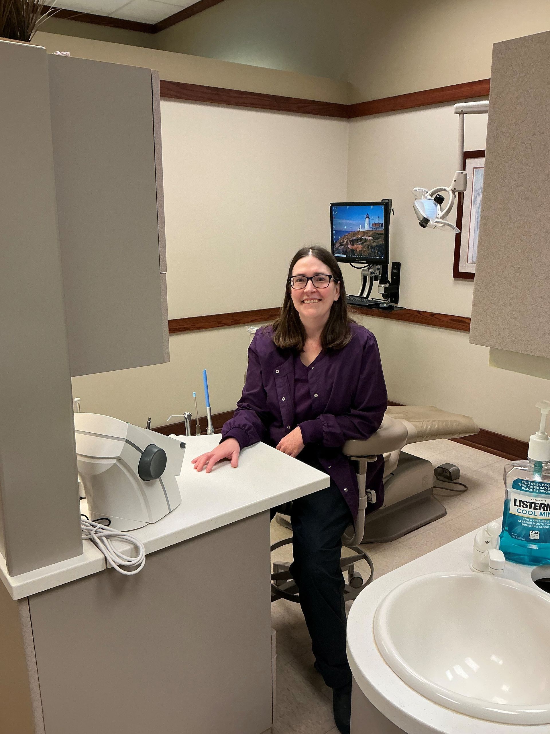Woman seated at a dental exam chair, smiling in a clinic room beside a sink and counter