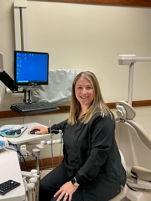 Dentist seated in exam room beside dental chair, smiling at camera with computer monitor in background