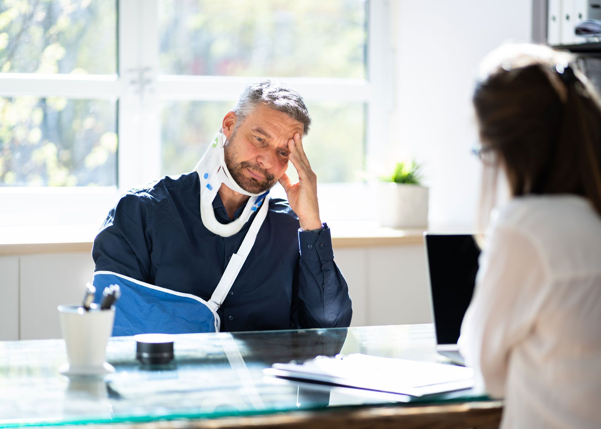 A man with a broken arm is sitting at a desk talking to a woman.