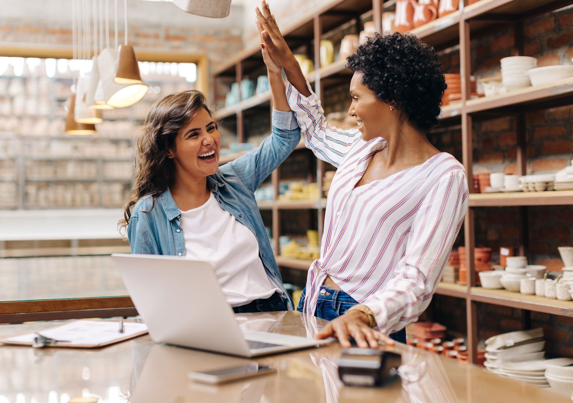 Two women are giving each other a high five while sitting at a table with a laptop.