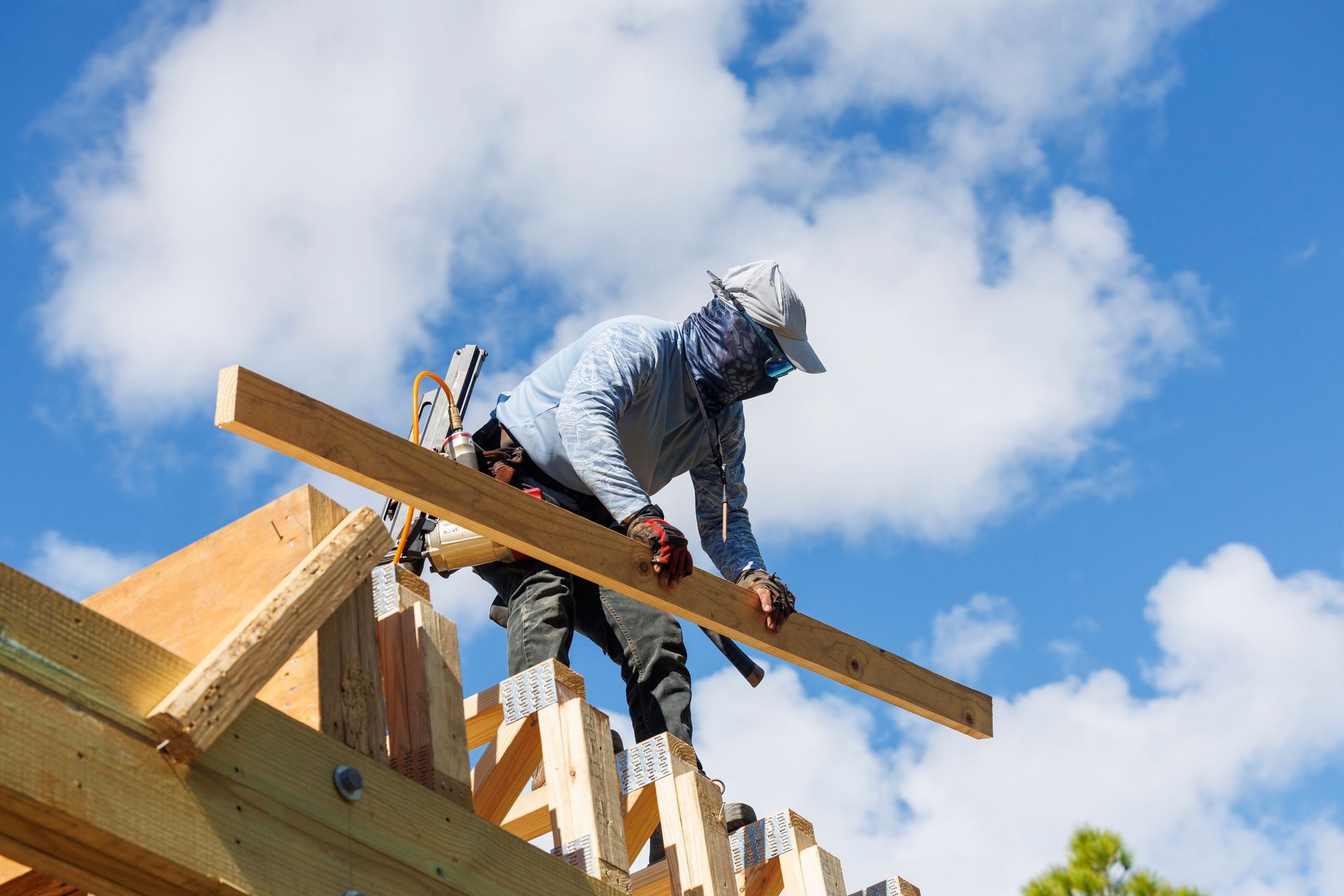 Worker secures a beam atop wooden roof trusses under a bright sky during construction.