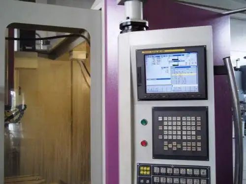 Pannello di controllo della macchina CNC con schermo che visualizza codice, pulsanti e sportello della macchina chiuso.