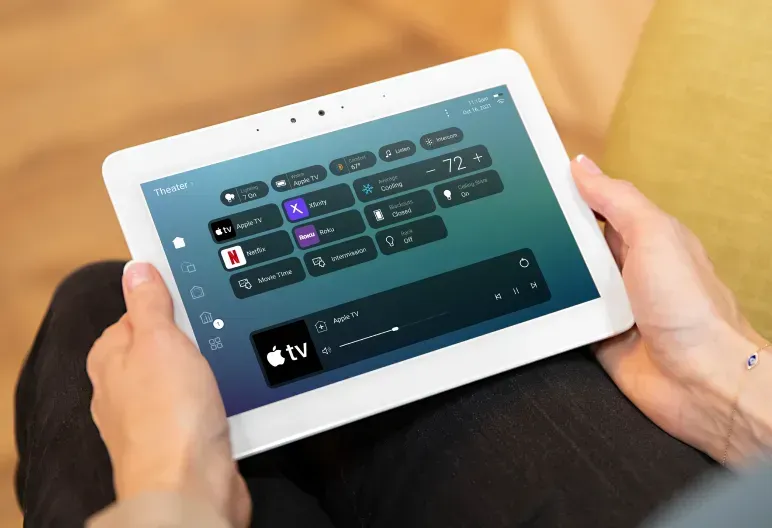 Person holding a tablet displaying a smart home control interface. The screen shows media controls and temperature settings.