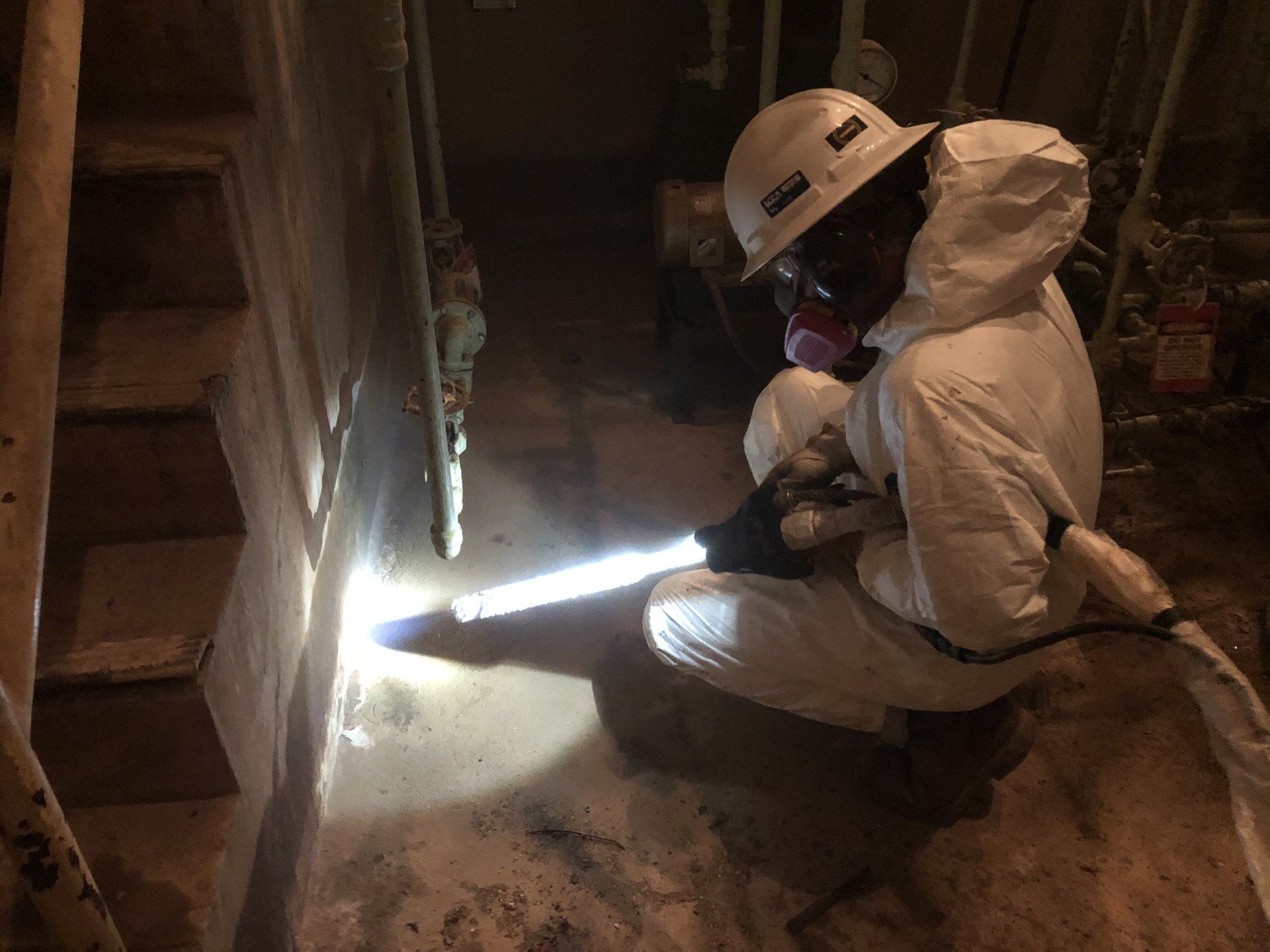 A man in a hard hat and protective suit is kneeling down in a dark room.