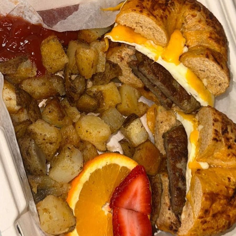 A bagel sandwich with eggs , sausage , cheese , potatoes and an orange slice.