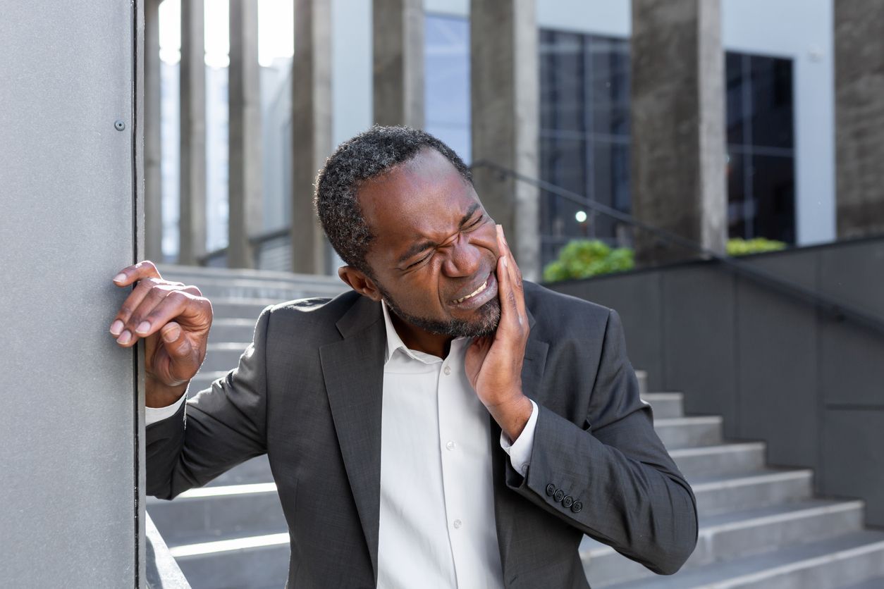 Man in suit grimaces, holding his jaw, leaning against a building.