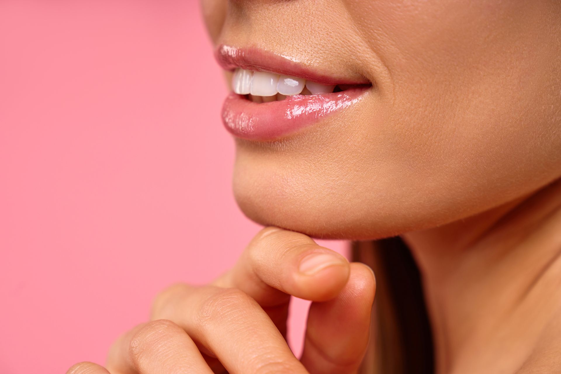 Close-up of a person's smiling mouth and chin, with hand touching chin. Pink lip gloss, pink background.