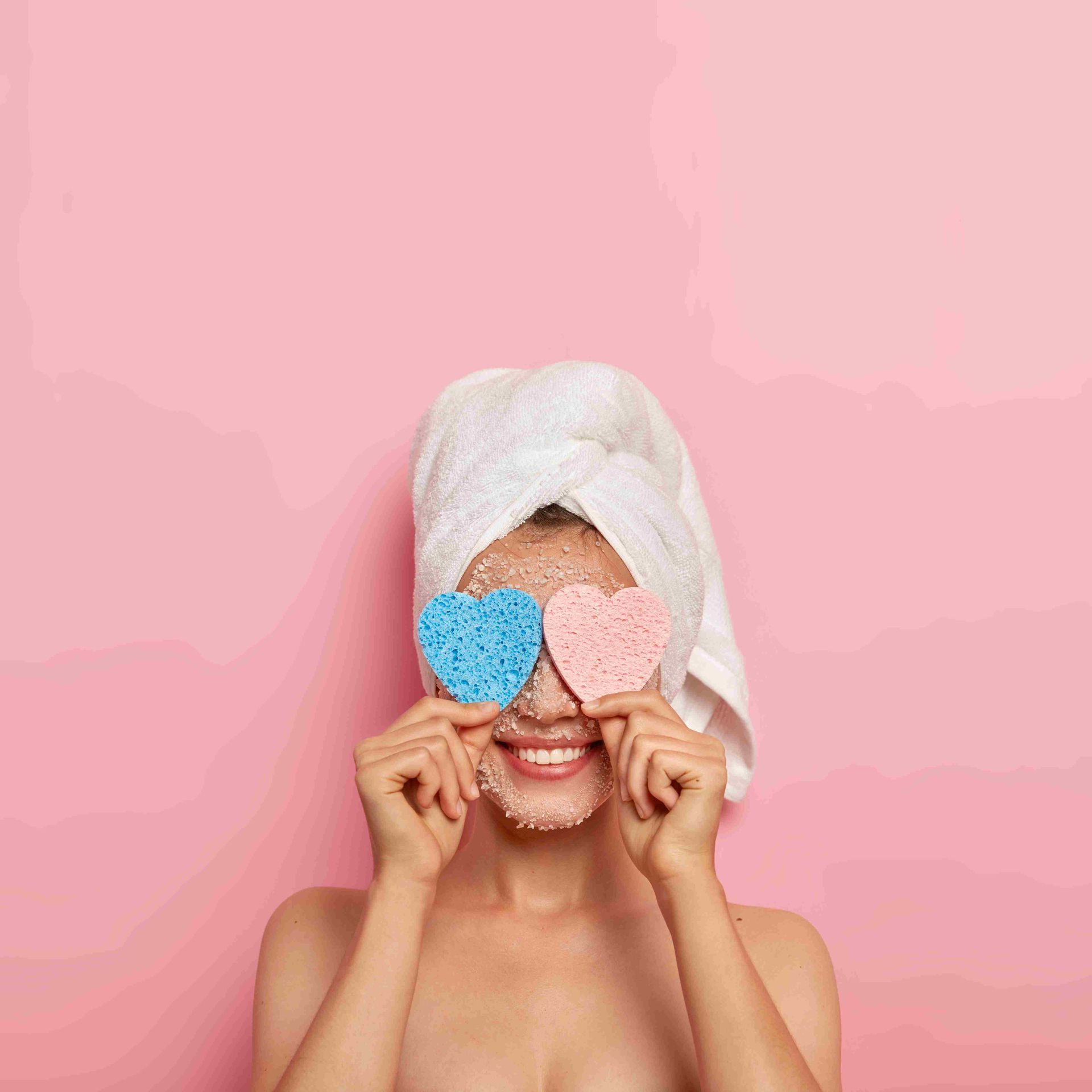 A woman with a pink background and towel on her hair holding heart shapes over her eyes