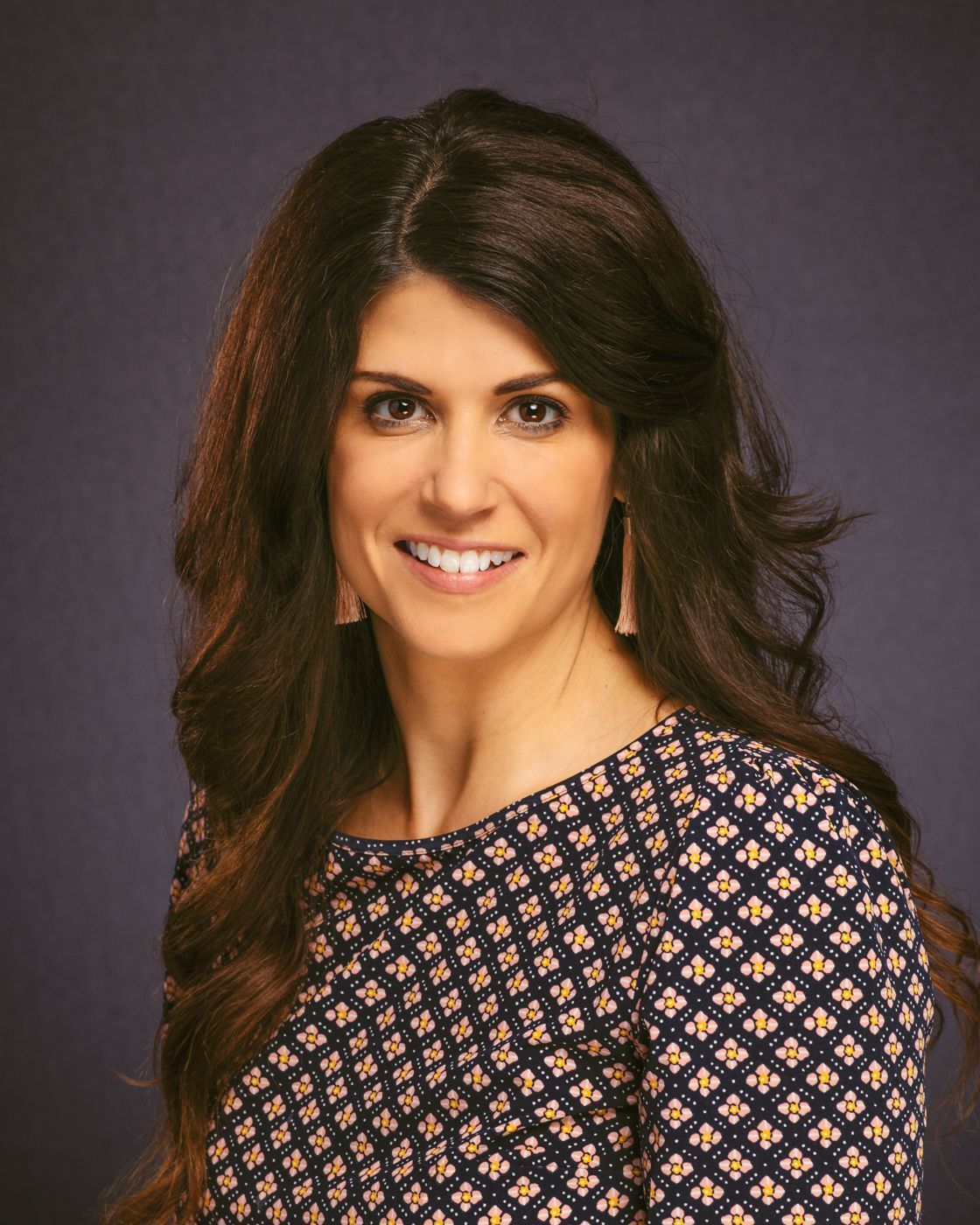 A woman with long hair is wearing a blue and gold dress and smiling for the camera.