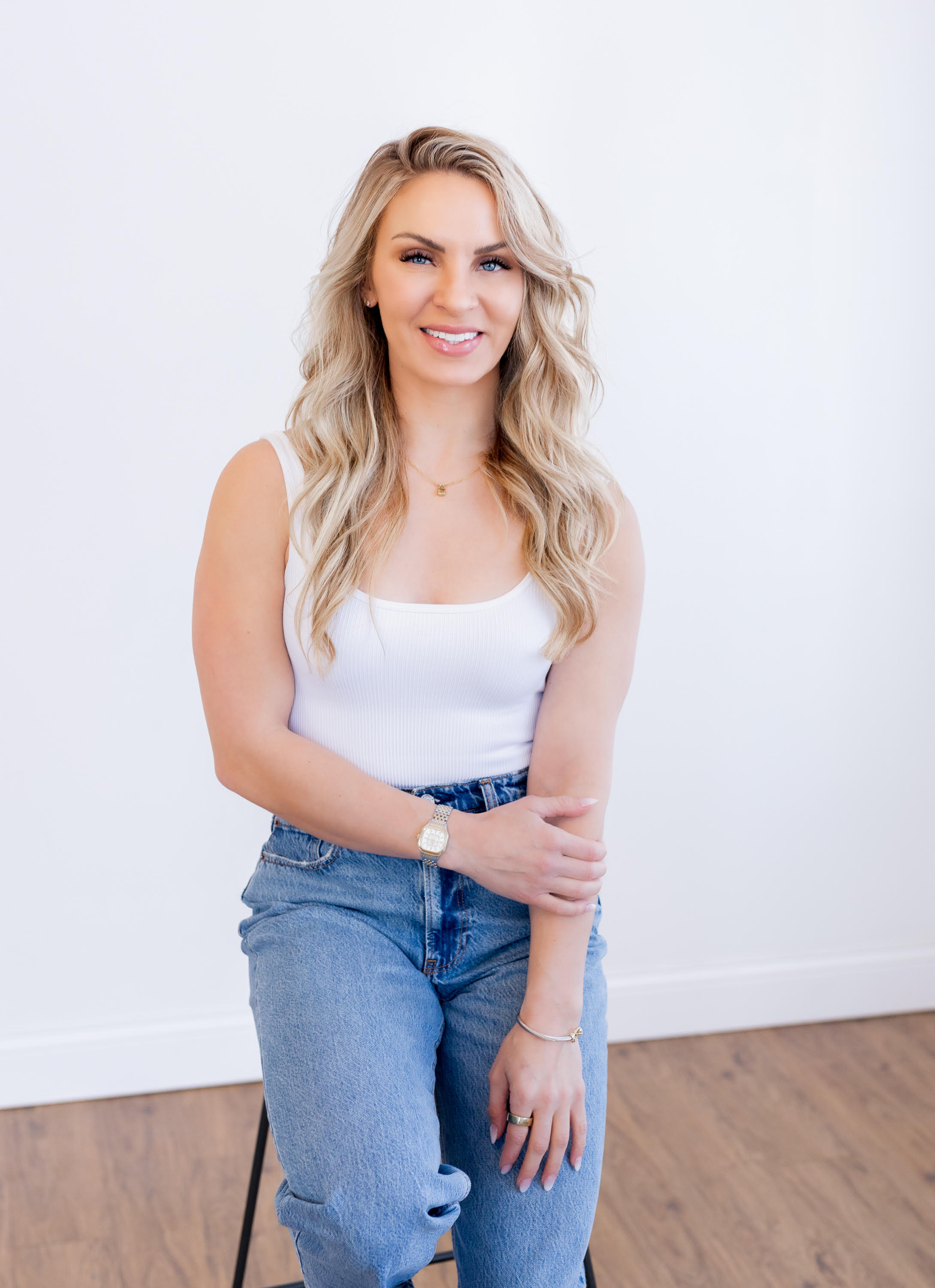 A woman in a white tank top and blue jeans is sitting on a stool.