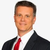 A woman in a suit is smiling in front of a wooden fence.