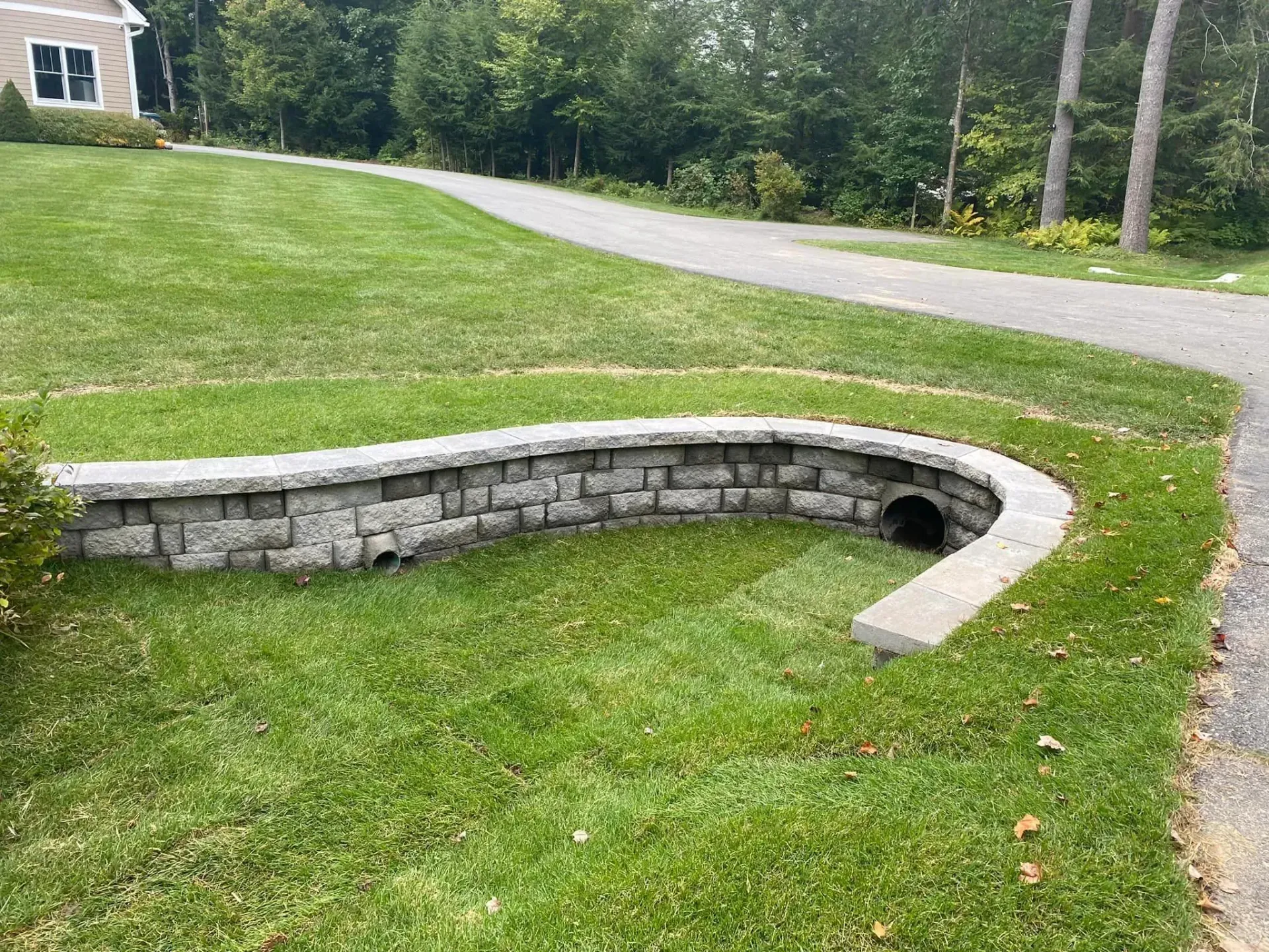 There is a drain in the grass next to a stone wall.