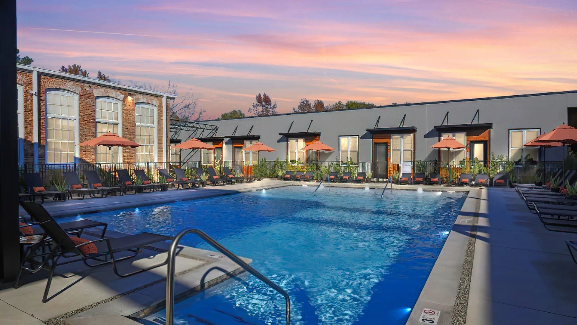 View of a swimming pool surrounded by lounge chairs and umbrellas at sunset.