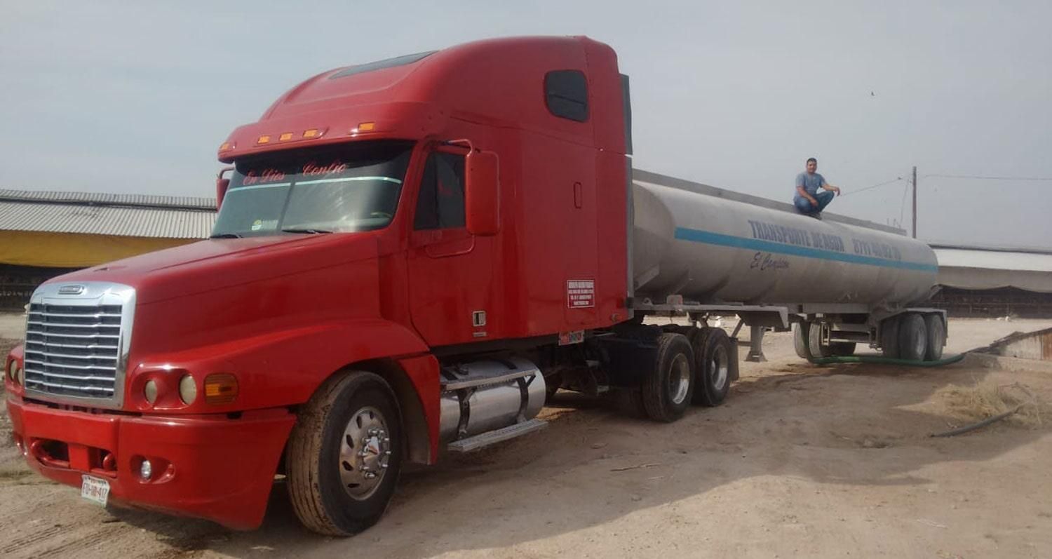 Un camión rojo con un remolque cisterna está estacionado en un terreno de tierra.