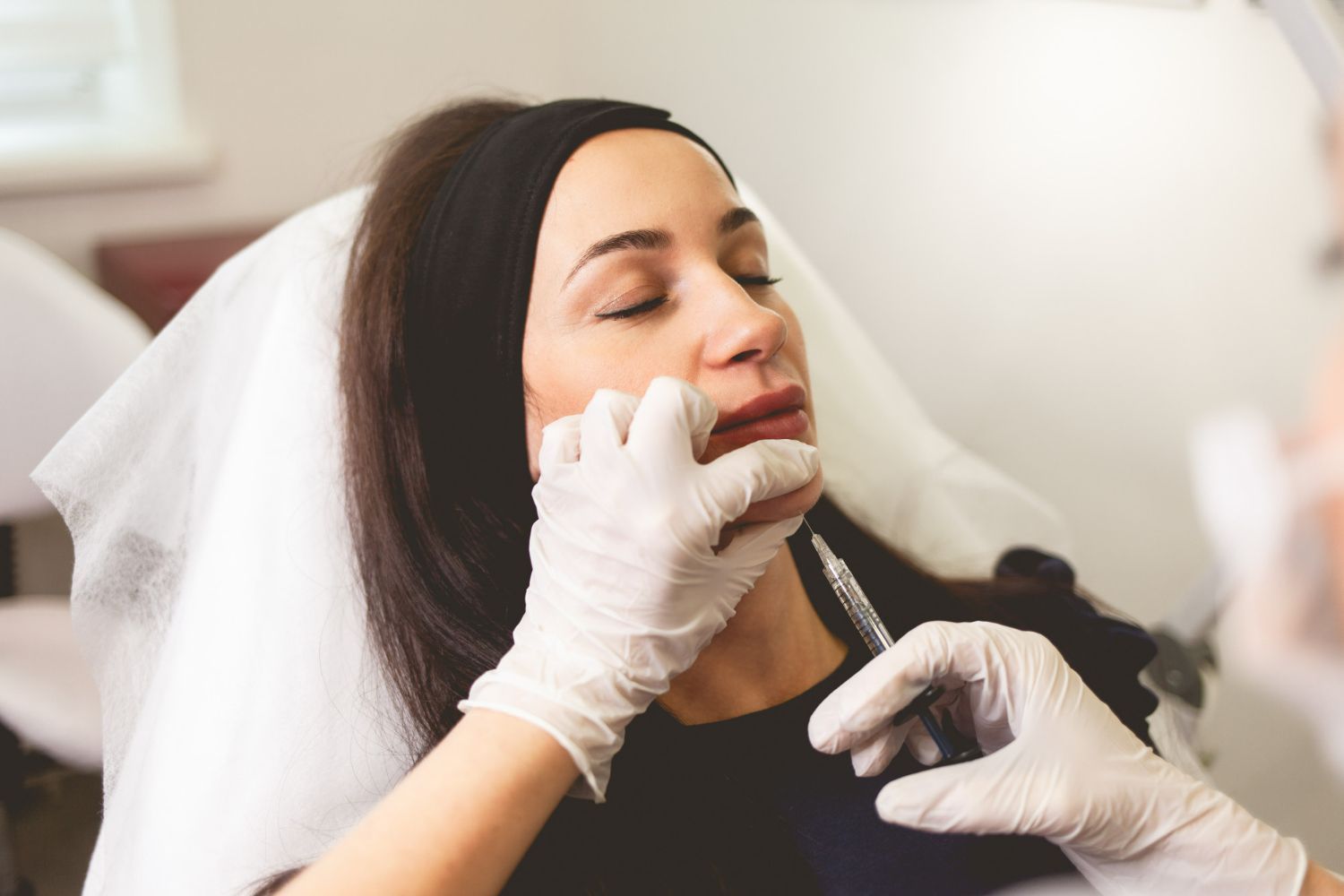 Woman receiving lip injections; doctor's gloved hands with syringe. She is reclining with eyes closed.