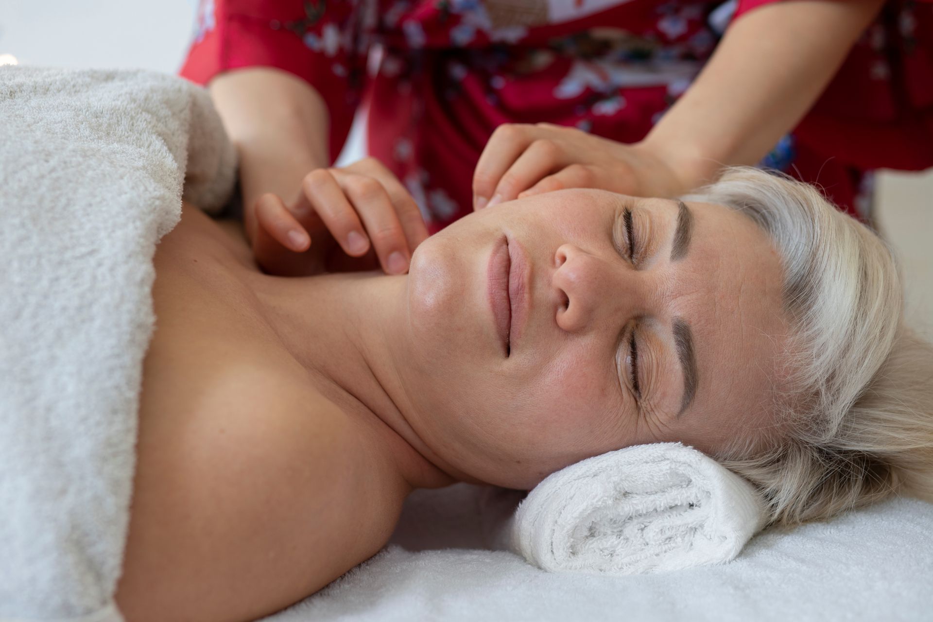 A person lies on a spa bed, eyes closed and relaxed, while a practitioner gives them a gentle facial massage.
