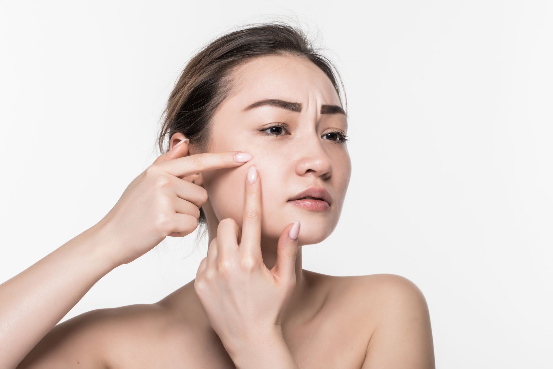 A person with a concerned expression uses their fingers to examine a blemish on their cheek against a white background.