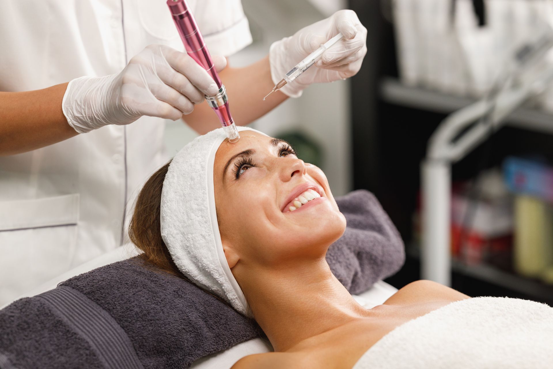 A person in a headband receives a microneedling treatment from a gloved practitioner using a pink electronic device.
