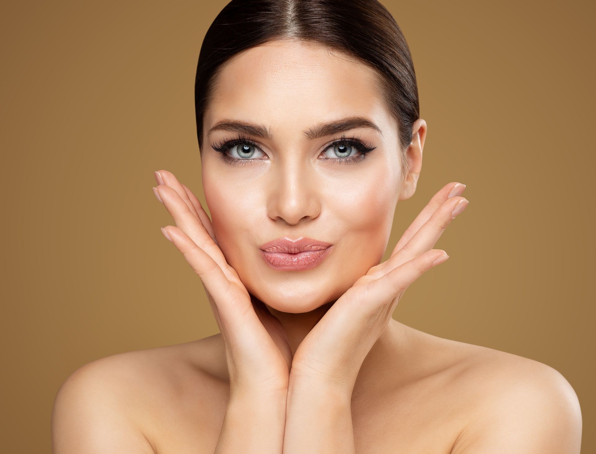 A person with smooth skin frames their face with both hands, looking toward the camera against a tan background.