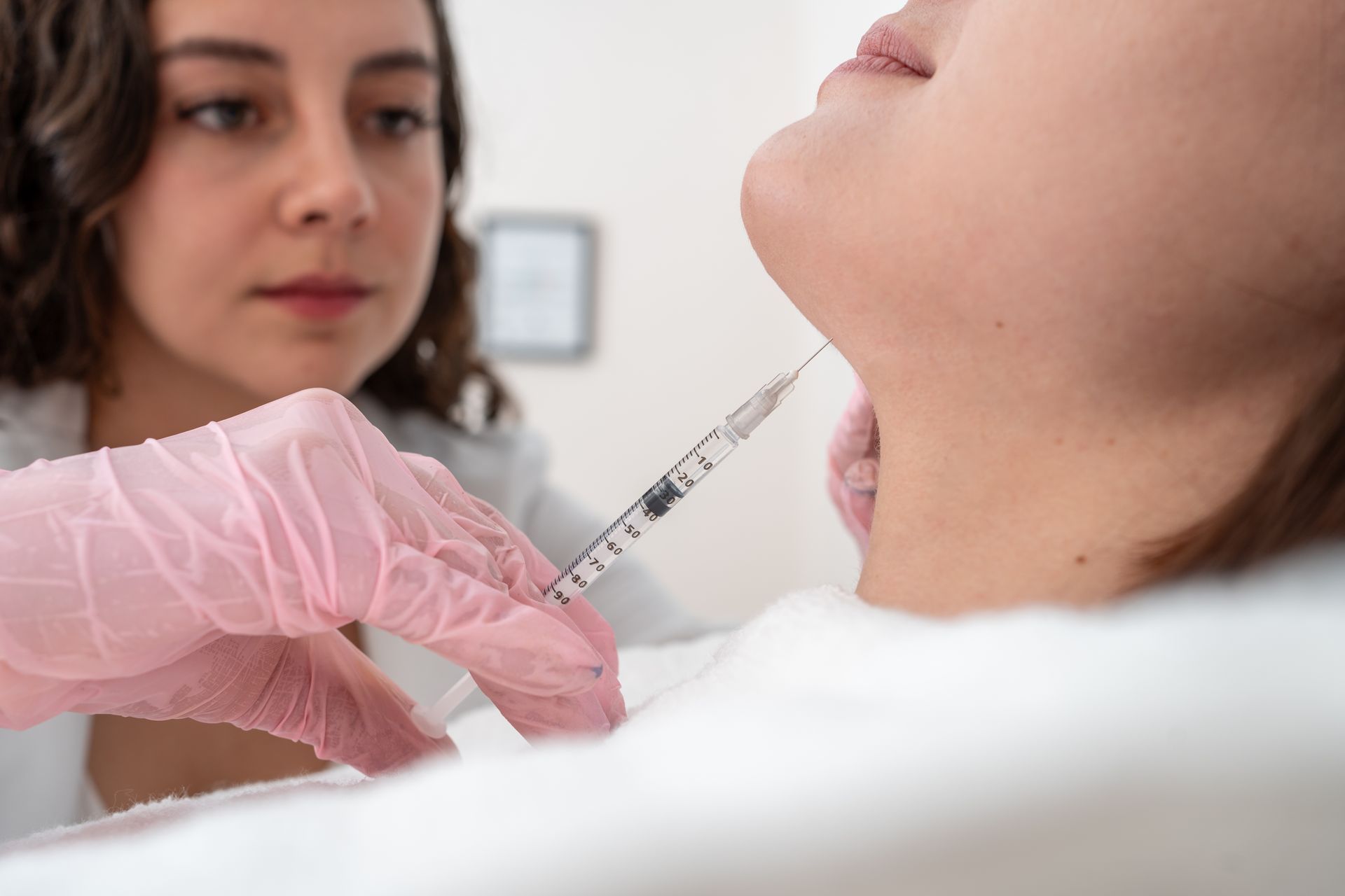 A practitioner in pink gloves administers a cosmetic injection into the chin area of a patient lying down.