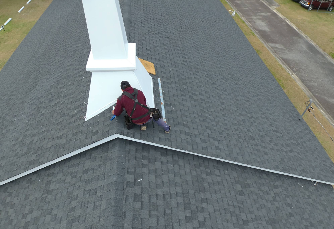 Two people on a gray shingle roof near a white chimney. One is working.