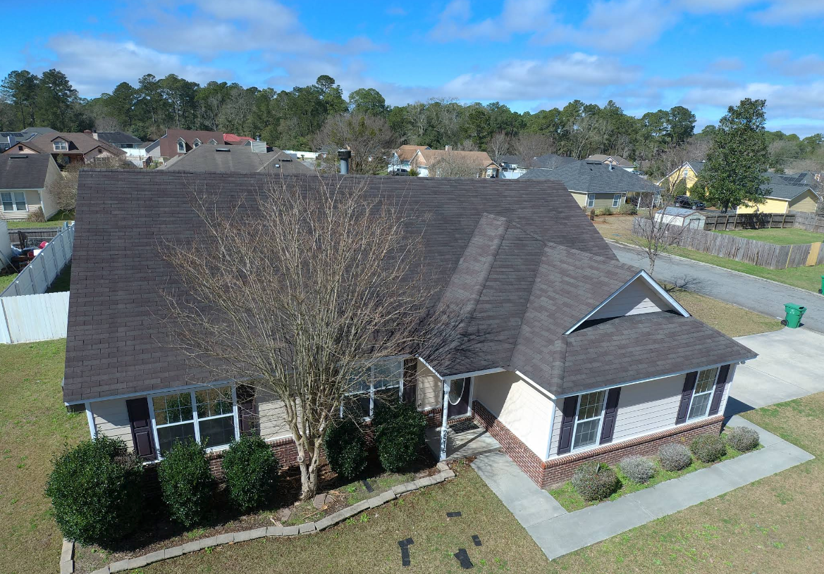 Aerial view of a suburban house with a dark roof and a tree in front. Blue sky and other houses are in the background.