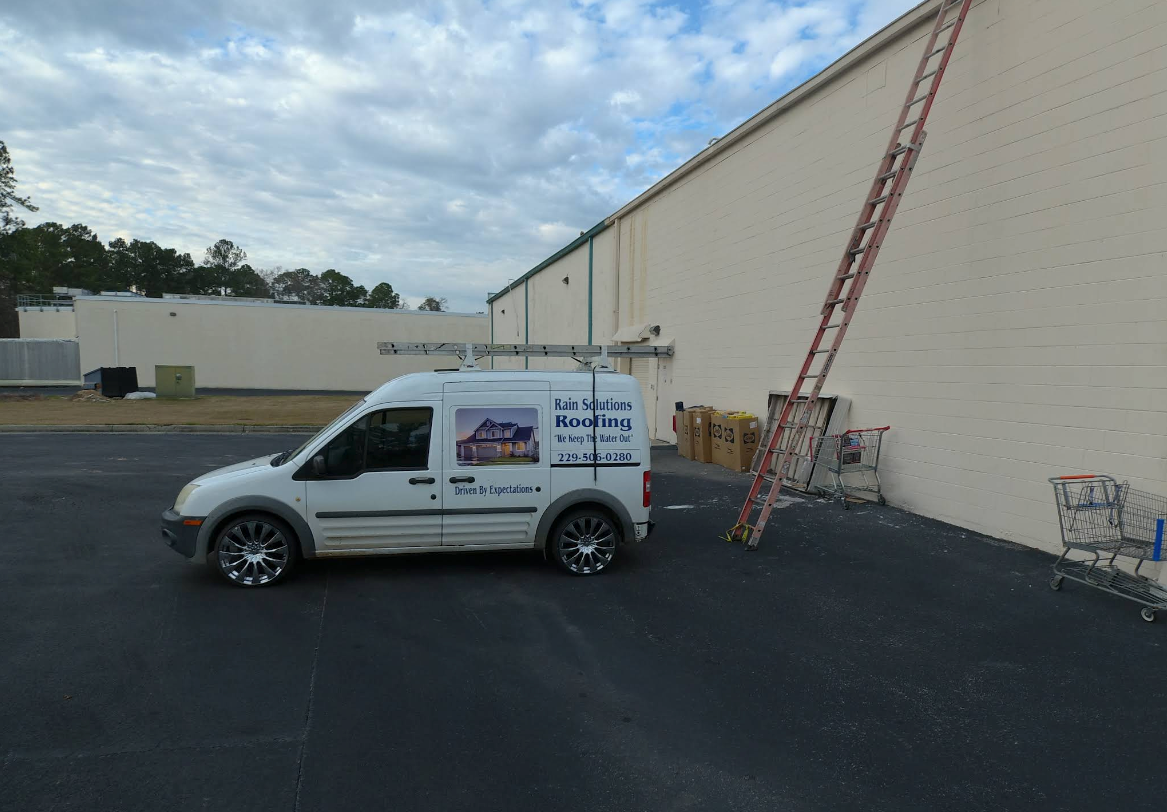 White van with business logo parked next to a large building. A ladder leans against the wall.