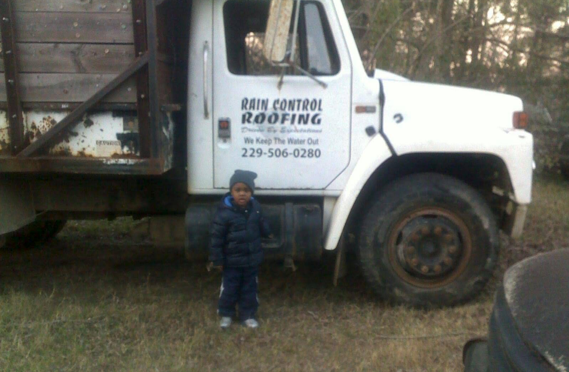 Child standing next to a white Rain Control Roofing truck with a wooden cargo bed; rural outdoor setting.