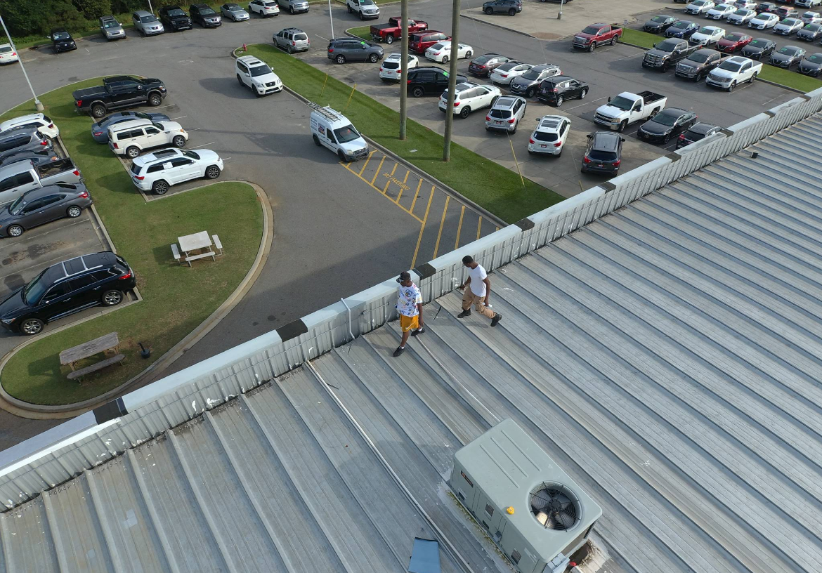 Two people on a metal roof, possibly repairing HVAC unit. Cars in a parking lot below.
