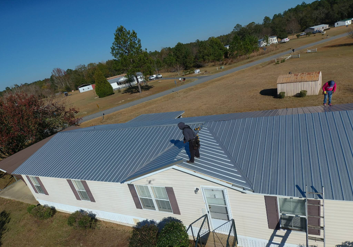Two people on a metal roof of a house, outdoors. Blue sky. Hills in background.