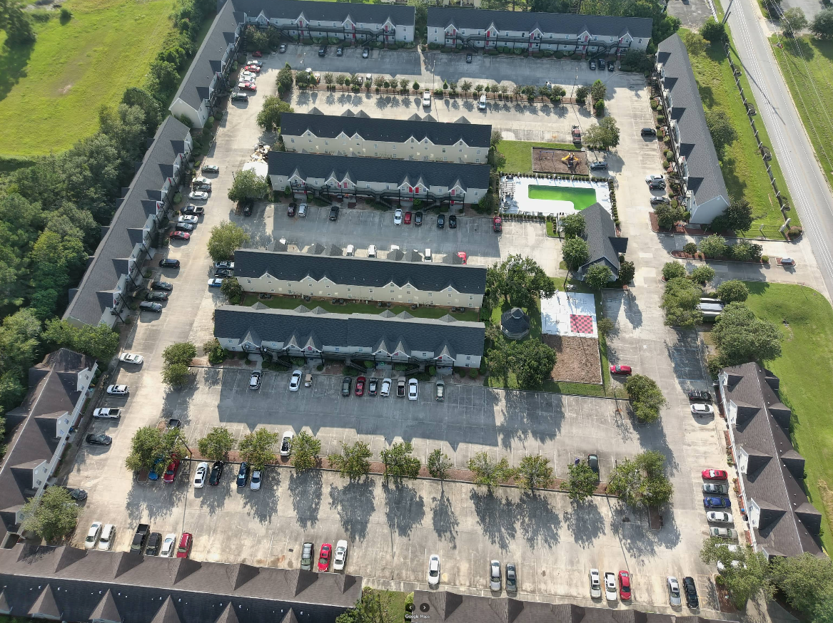 Aerial view of a residential complex with townhouses, parking areas, and a small pool surrounded by trees.