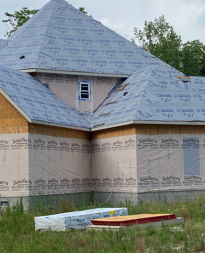 A partially constructed house with blue roof sheathing and light brown siding, set in a grassy area.