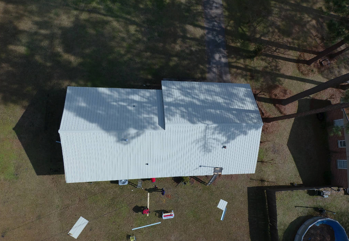 Overhead view of a house with a white metal roof. Equipment and materials are scattered on the lawn.