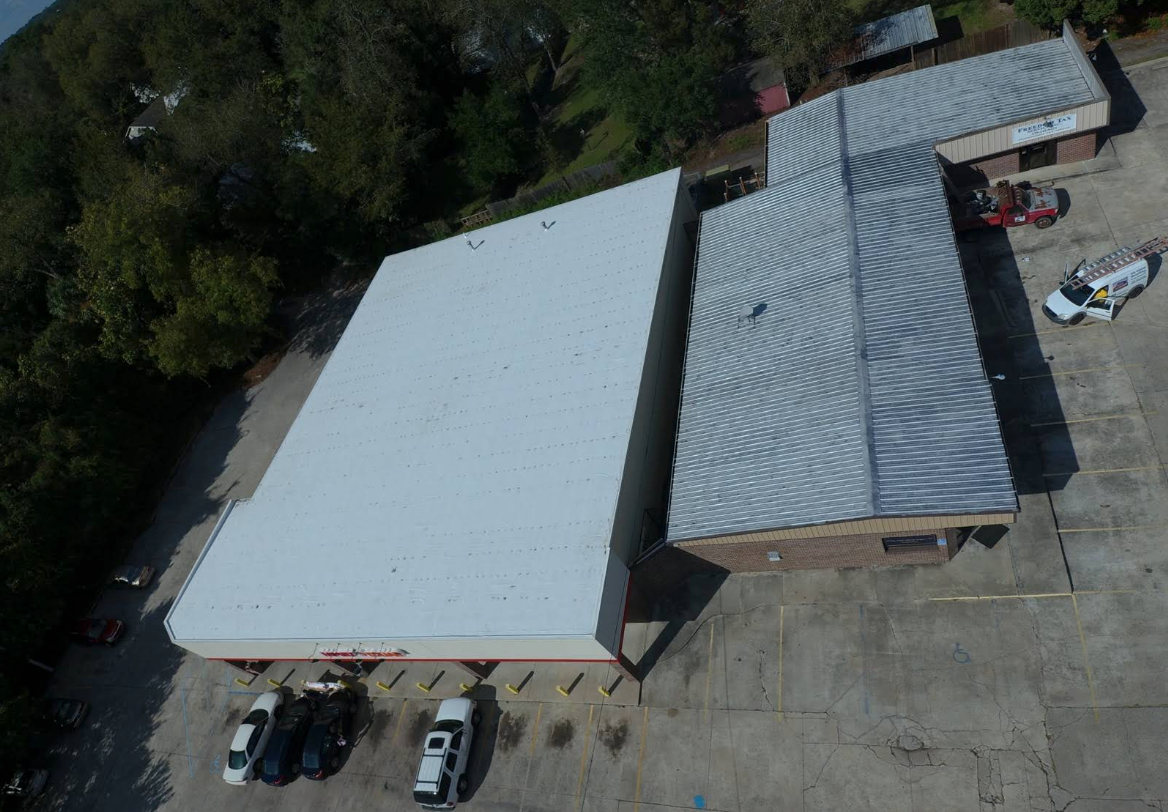 Overhead view of a commercial building with white and corrugated metal roofs, surrounded by a parking lot and trees.