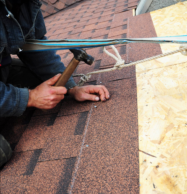 Person hammering roofing shingle on a roof, safety harness visible.