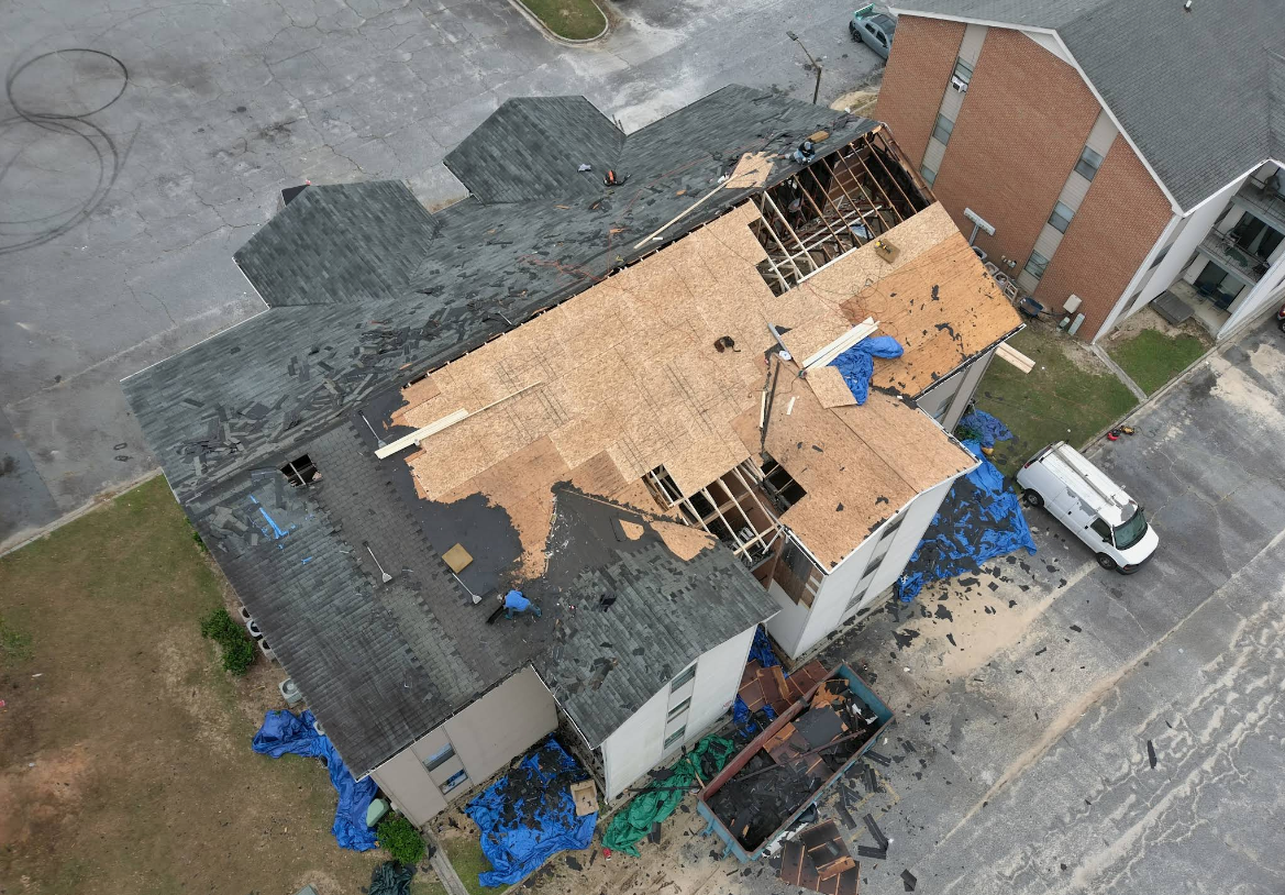 Aerial view of damaged apartment building roof after a storm, with missing shingles, exposed structure, and blue tarps.