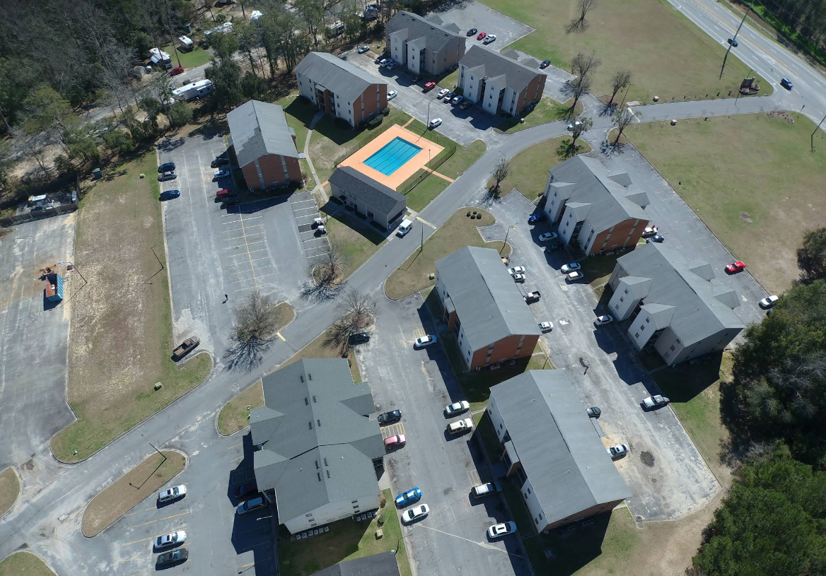 Aerial view of apartment complex with brick buildings, a pool, parking areas, and a road.