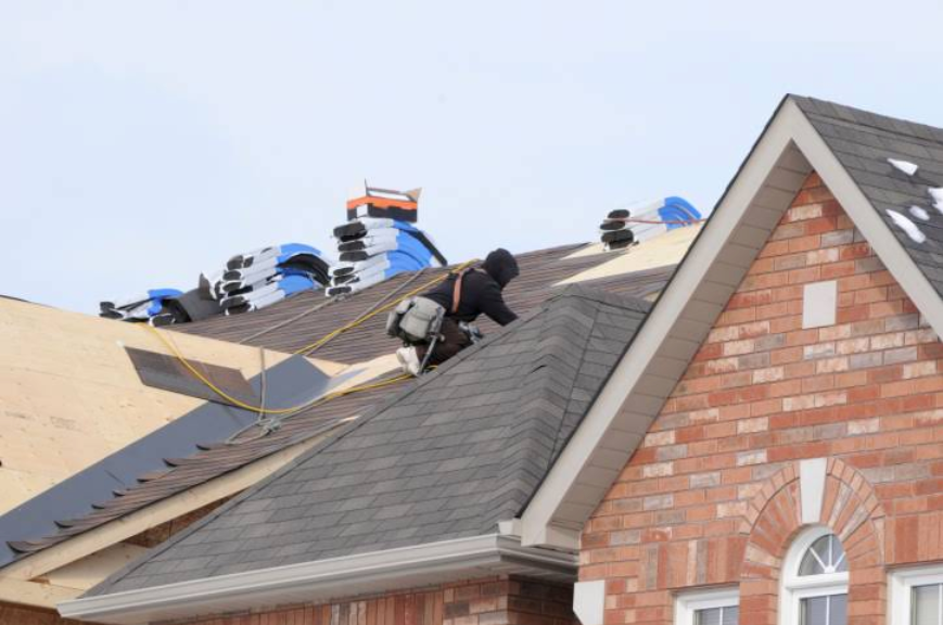 Roofer installing shingles on a house roof; other roof sections are exposed.