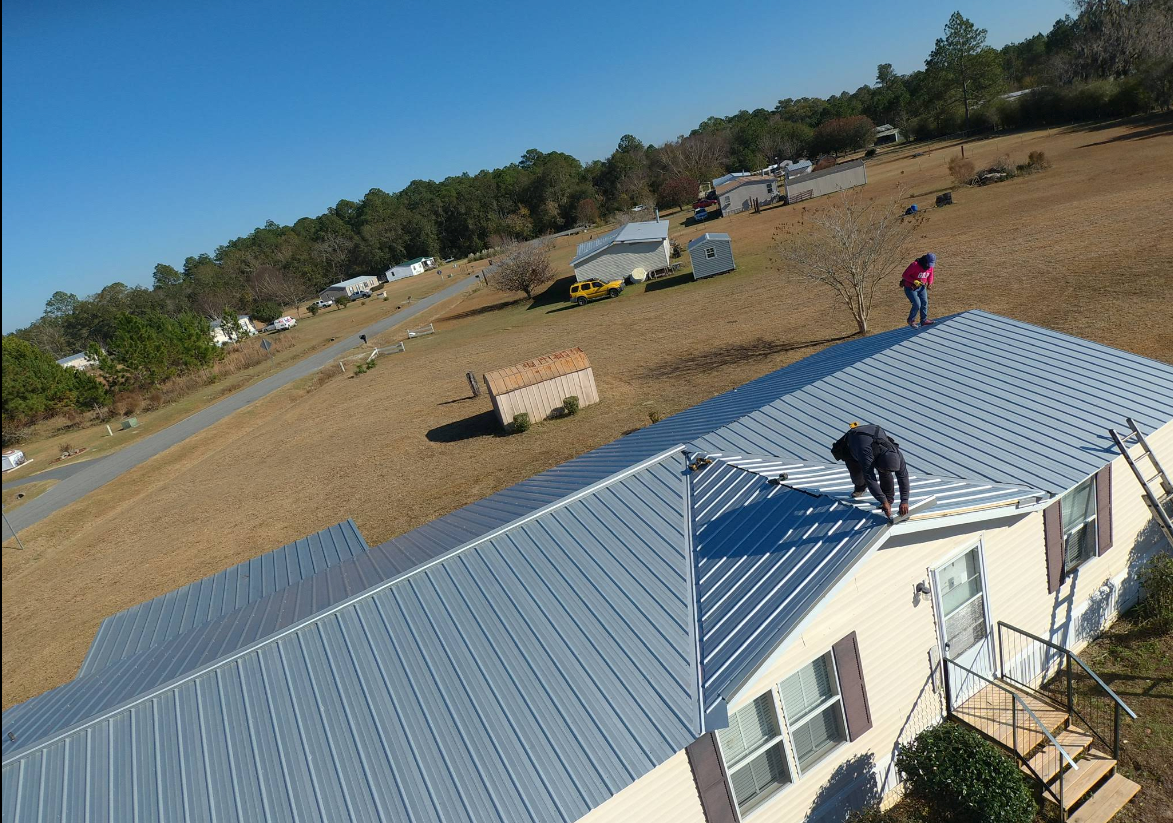 Two people on a metal roof, working. A ladder is leaning against the side of the house. Trees and a hill are in the background.