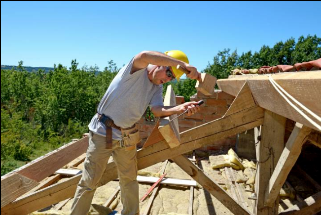 Carpenter on a roof, hammering wood. Wearing a hard hat and safety glasses, sunny day, surrounded by trees.