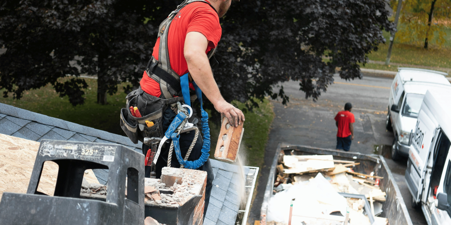 Roofer in safety harness removes debris from chimney, placing it in a dumpster. Second worker nearby.