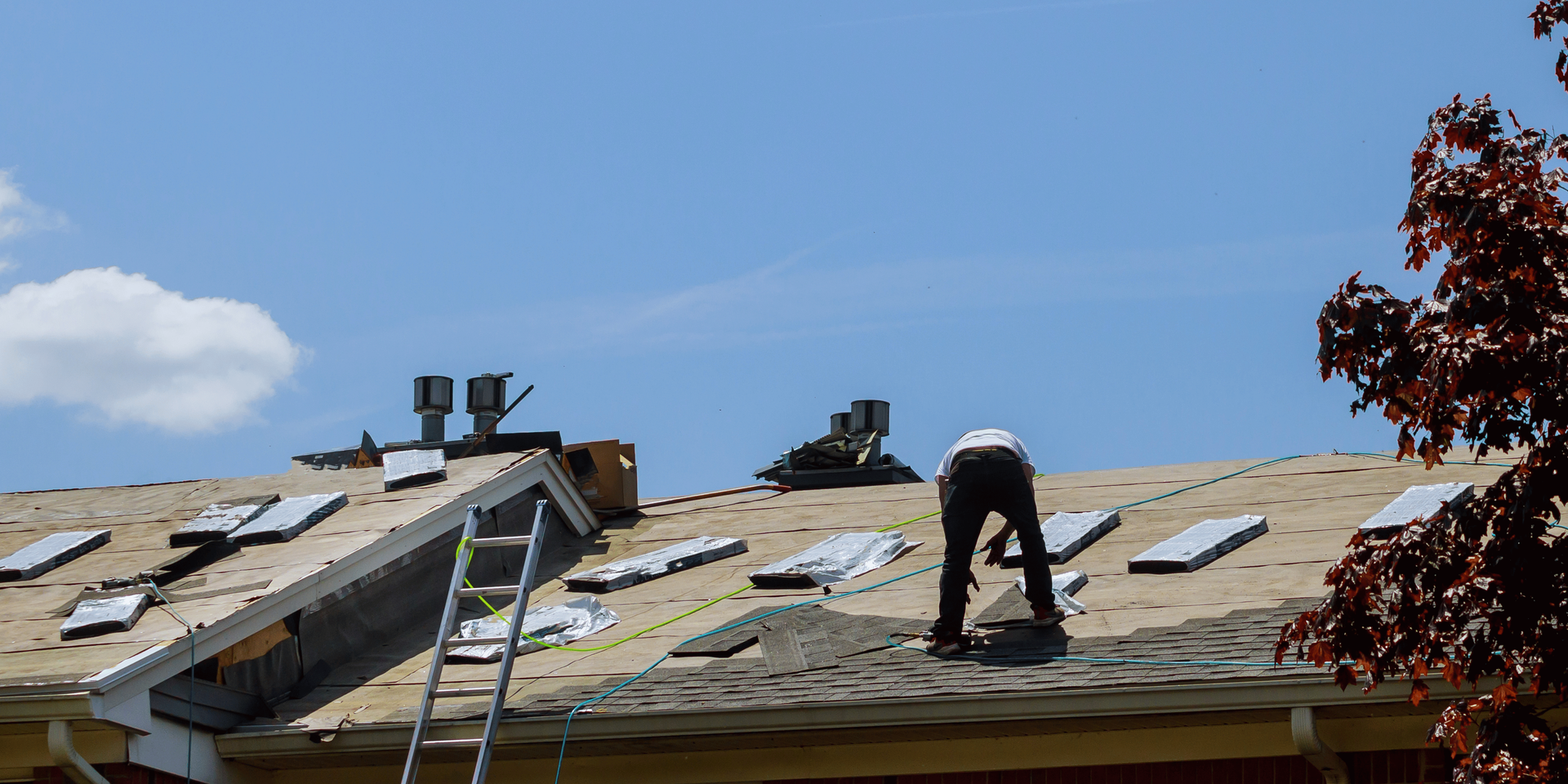 Person working on a roof, removing old shingles. A ladder is present. Blue sky.