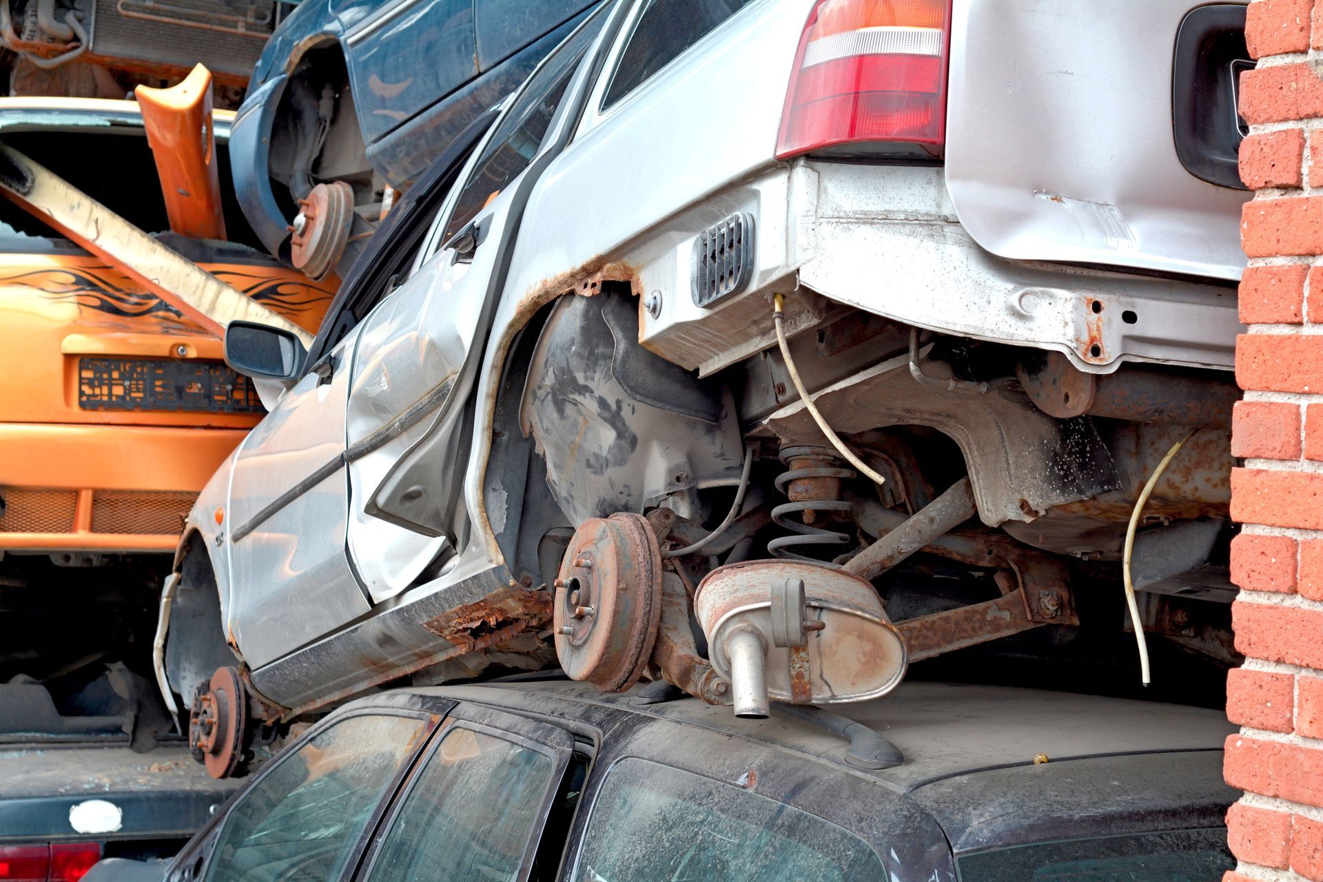 Una carrozzeria argentata, priva di ruote e parti, è accatastata sopra altri veicoli in una discarica, accanto a un muro di mattoni.