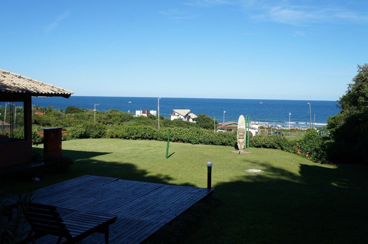 Gramado verdejante com vista para o oceano azul sob um céu claro, pequenas estruturas à distância.