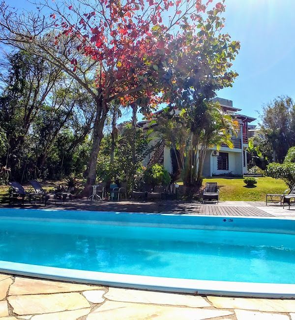 Piscina com água azul, gramado verdejante e uma casa com árvores altas ao fundo.