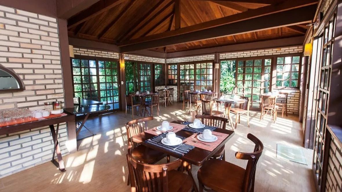 Sala de jantar ensolarada com mesas e cadeiras de madeira; paredes de tijolos e janelas; vista para a floresta.