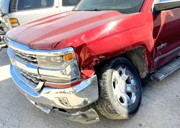 Red pickup truck with front-end damage, crumpled fender, and scraped bumper in a parking lot