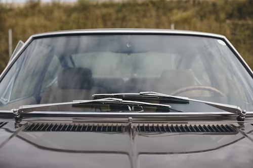 Close-up of a dark car's windshield with wiper blades, hood vents, and interior visible.