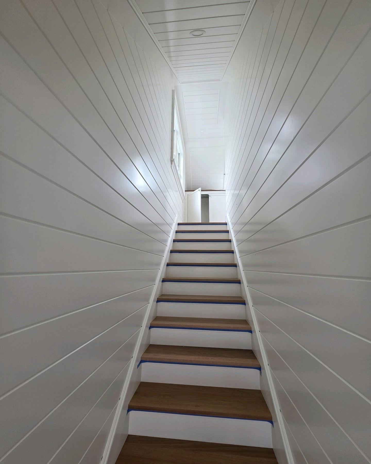 Narrow white staircase rising into a bright hallway with slanted walls and wooden steps